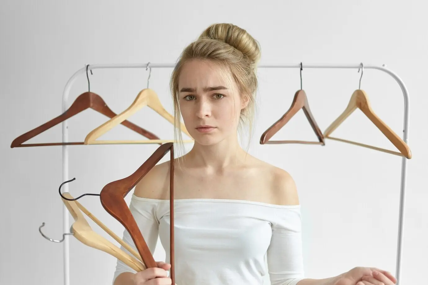 Foto de una joven caucásica frustrada, con un top blanco de hombros descubiertos, posando en un dormitorio, sosteniendo perchas vacías en las manos y con una barra detrás de ella, perpleja después de que su esposo la dejara llevándose toda su ropa.