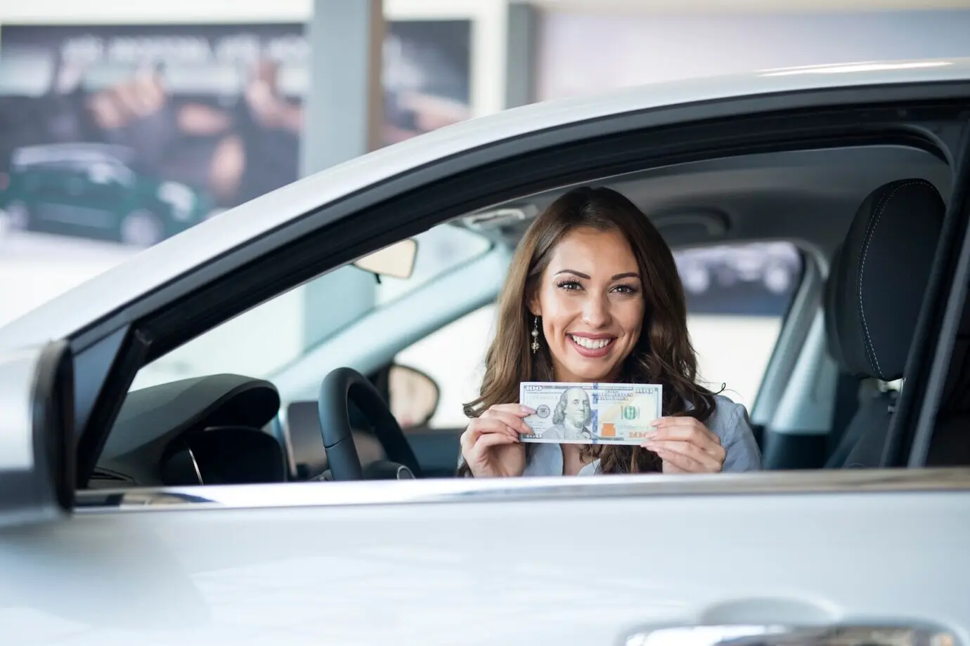 Mujer alegre sentada en el coche nuevo, sosteniendo un billete de dólar estadounidense.